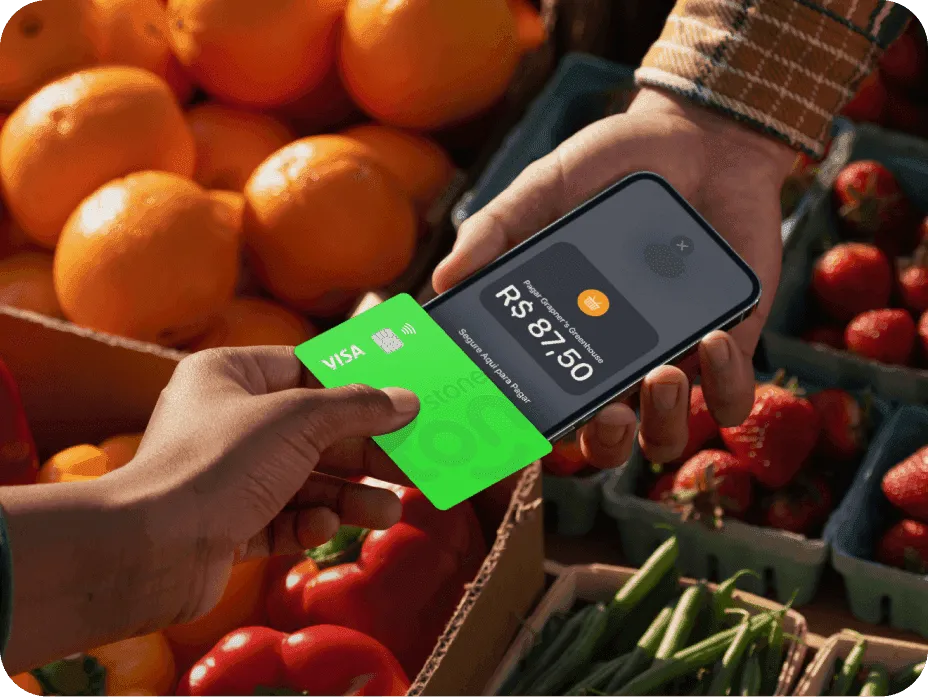 Pessoa fazendo pagamento com cartão Visa verde da Super Conta Ton em uma feira ao ar livre, usando o sistema de aproximação. A tela do dispositivo mostra o valor de R$ 87,50.