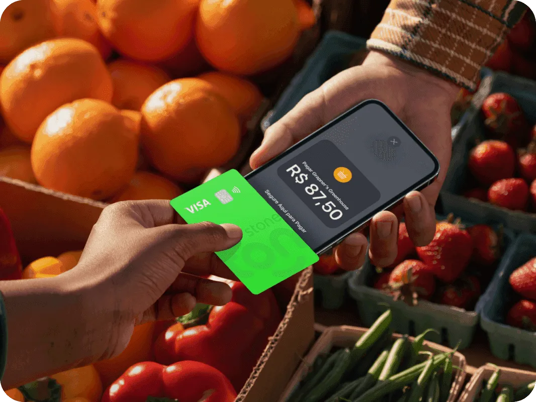 Pessoa fazendo pagamento com cartão Visa verde da Super Conta Ton em uma feira ao ar livre, usando o sistema de aproximação. A tela do dispositivo mostra o valor de R$ 87,50.
